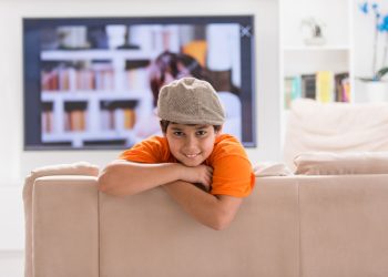 Dziecko siedzi samotnie w salonie z zegarem na ścianie i zamkniętymi drzwiami wejściowymi